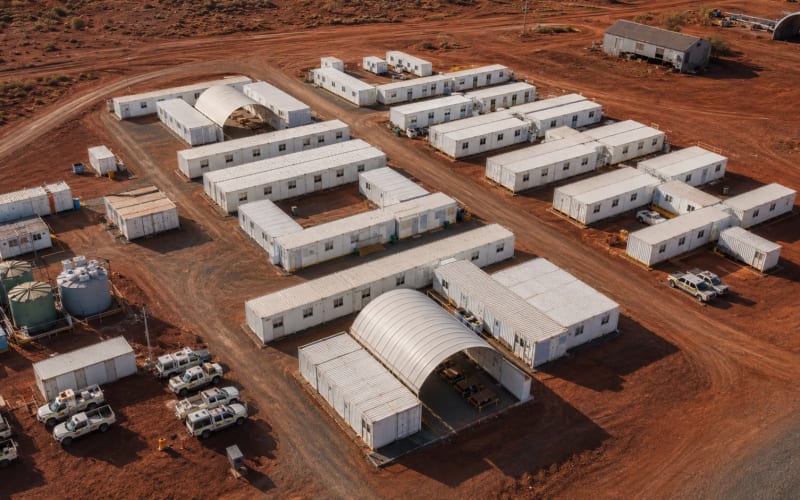 Aerial view of a modular mining camp set up on red dirt: rows of white accommodation and amenity buildings, two curved-roof shelters, bulk water tanks, and vehicle access roads.