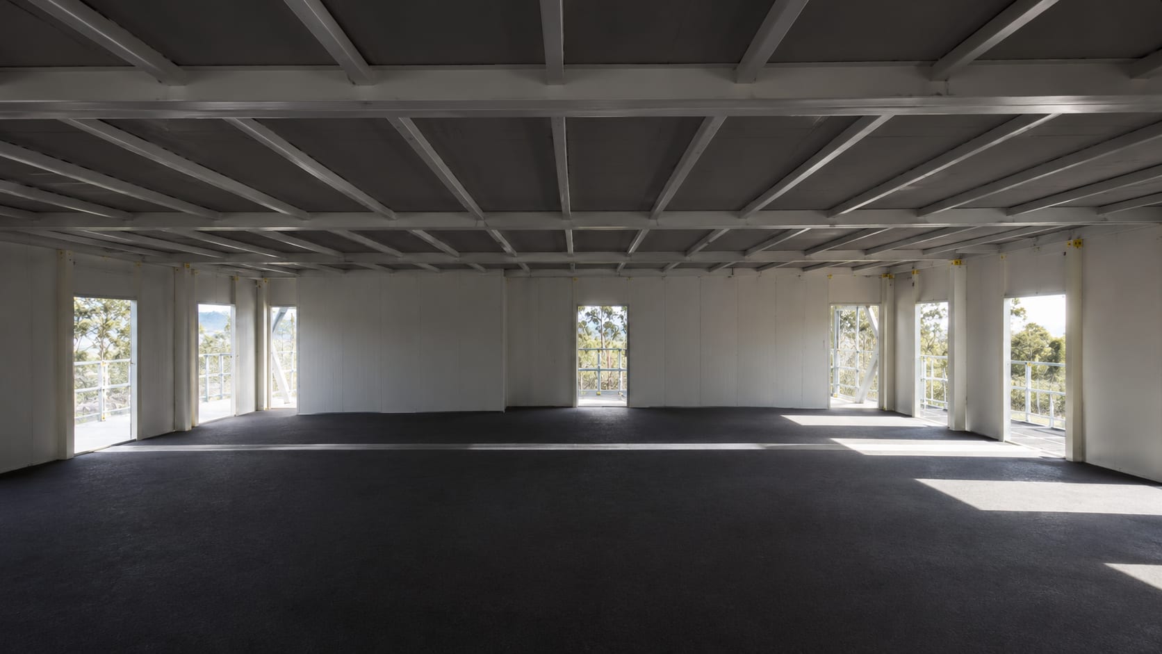 Interior of the Australian Federal Police modular training facility, a large open-plan floor with the white-painted modular steel frame exposed overhead, charcoal carpet, and perimeter windows onto bushland.