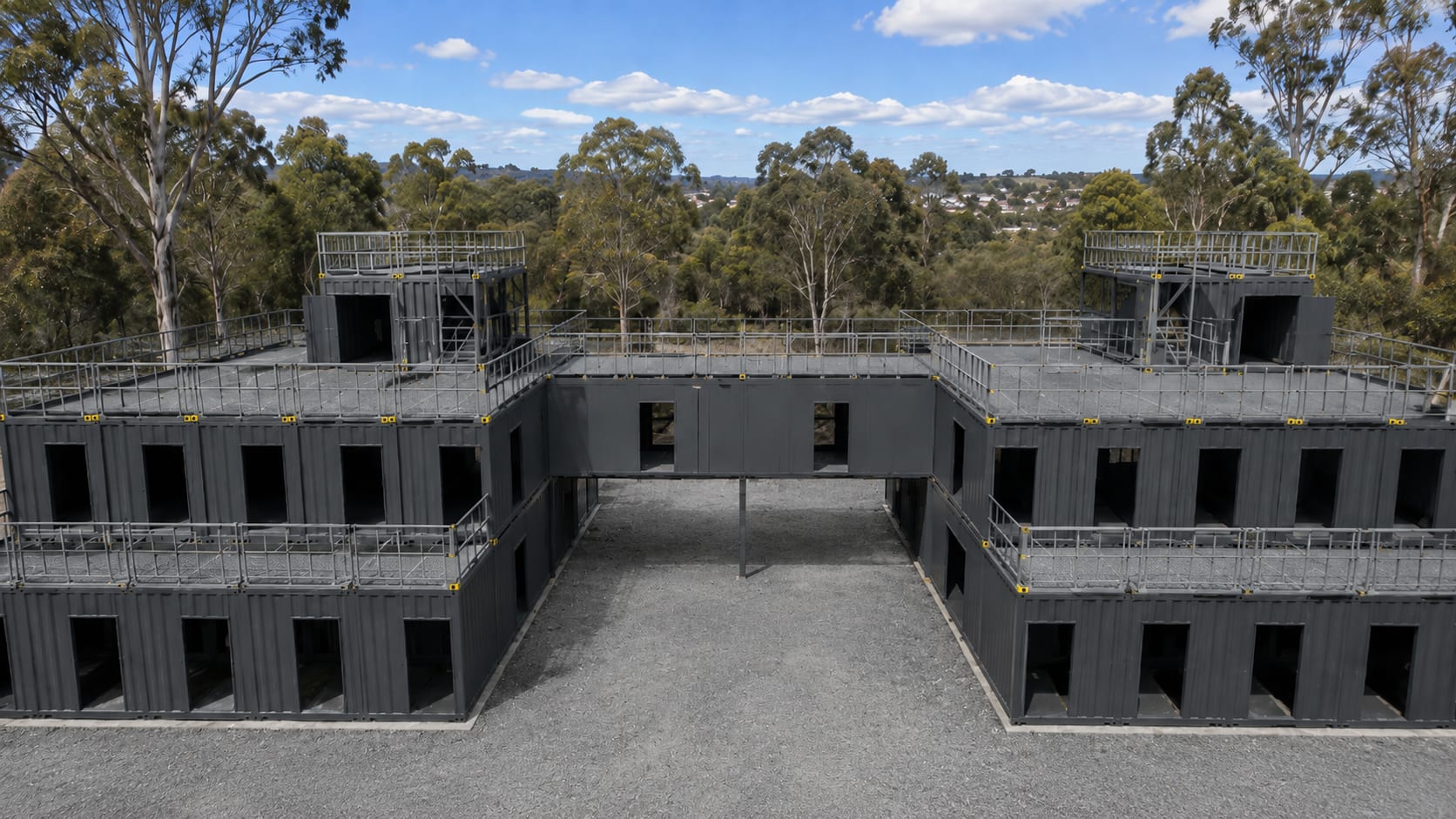 Aerial front view of the Australian Federal Police modular training facility, two wings of charcoal container modules around a central courtyard joined by an elevated link, with rooftop balustrades, in Australian bushland.