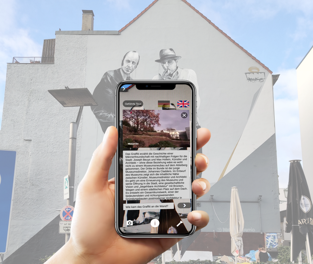 Eine Hand hält ein Smartphone und scannt ein Mural von Beuys. Auf dem Display werden Informationen als Text und Bild zu Beuys angezeigt.