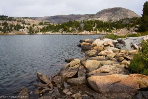 A rocky shore along an alpine lake with trees and mountains across the lake 