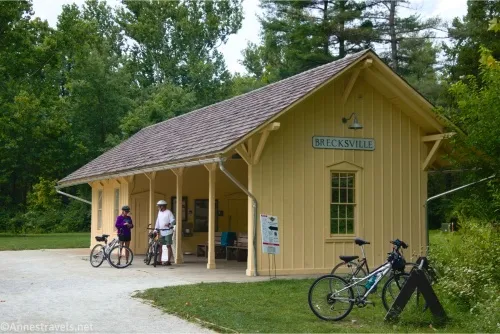 Two bicycles and two bicyclists stand beside the yellow building of Brecksville Station 