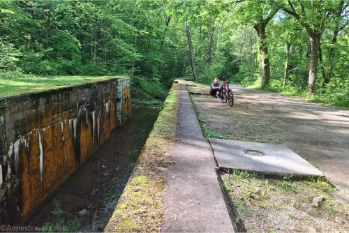 An old canal lock with orange walls, a path, a bicyclists, and green trees 