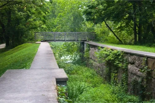 A wooden bridge crosses the end of an old canal lock filled with green plants 