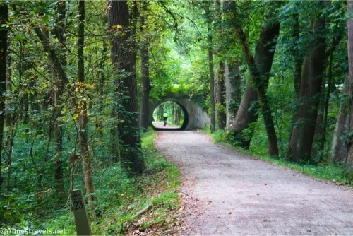 A wide path travels through the woods to a tunnel through which a bicyclist in a yellow shirt has just ridden 