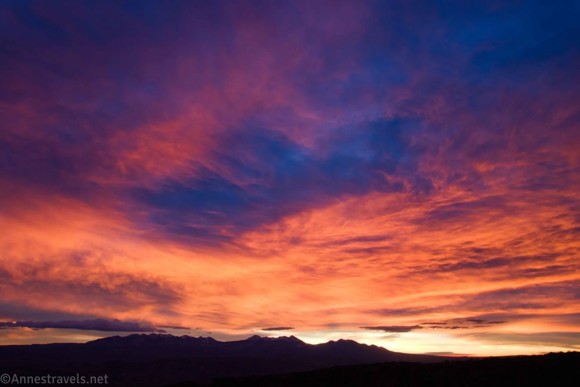 A sunrise with blue, purple, pink, orange, and yellow clouds over a dark, silhouetted mountain range