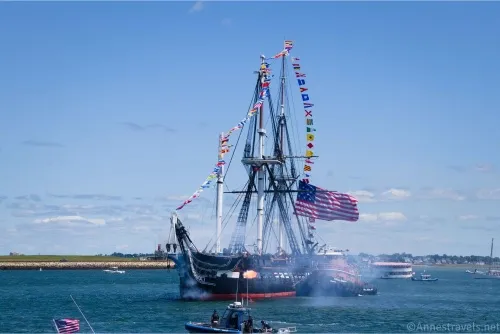 A historic warship fires a cannon at the shore with flags flying