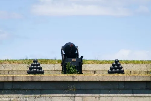 A cannon and two piles of cannonballs on top of a stone shelf with blue sky and clouds in the background