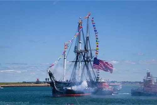 The historic warship USS Constitution fires a cannon with flying flags and two tugboats close by