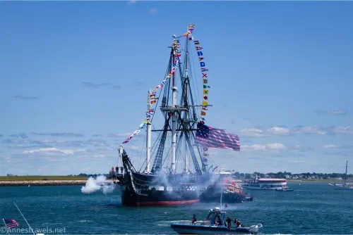 A historic warship with flying flags produces smoke with cannon fire in the blue ocean