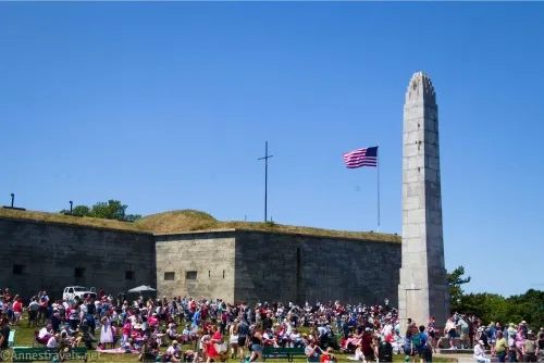 People sit on green grass beside a stone monument and below the stone walls of a fort with an American flag flying on top of the wall