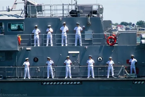 People in white Navy uniforms stand on two decks of a small gray boat