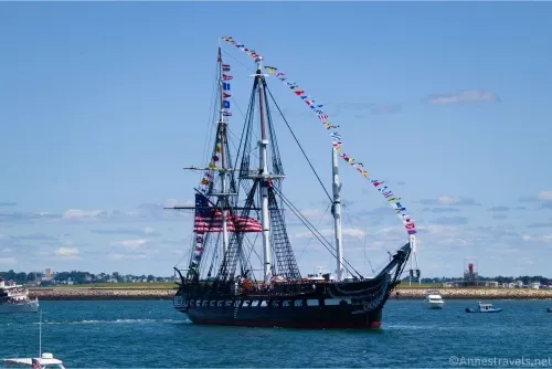 A historic gunboat with flying flags sails on blue water with clouds and blue sky in the background