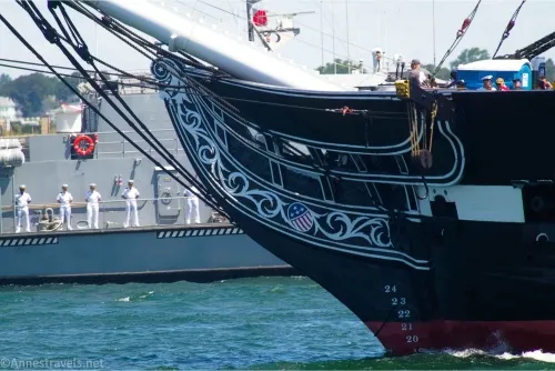 Prow and hull of a warship with fancy decorations and people looking over the railing