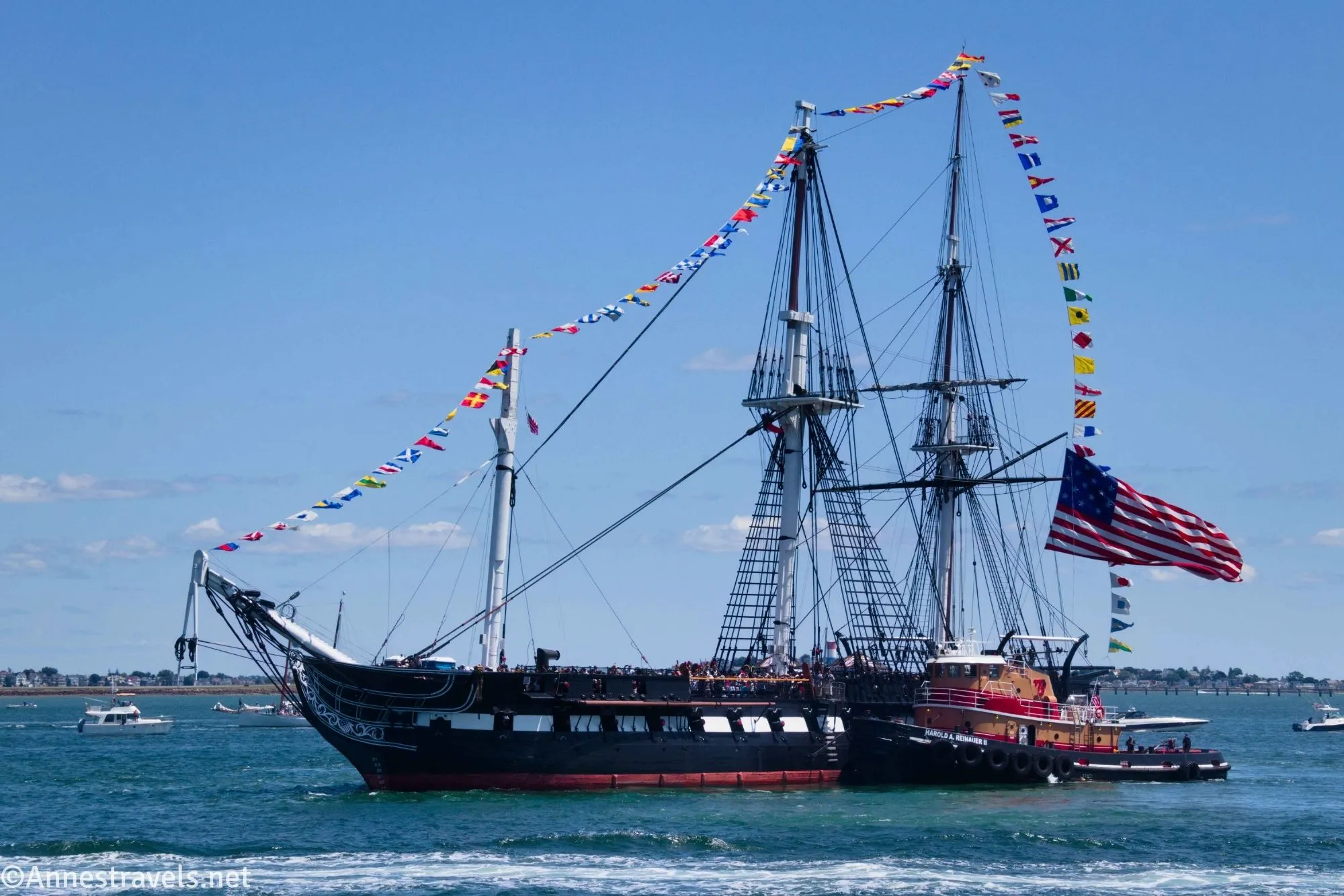 The USS Constitution warship with flags flying and a tug boat sail back toward Boston Harbor