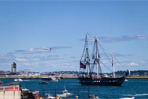 An airplane flies above Boston Harbor as the USS Constitution and other boats sail in the water