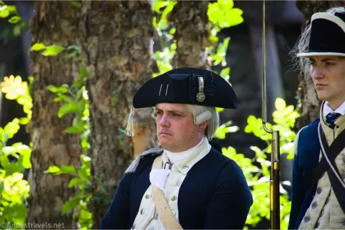 A man in front of a tree wears a tri-cornered hat, a wig, and Revolutionary War-era clothing
