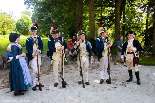 Four soldiers and a woman in period costume wait on a gravel path with trees in the background