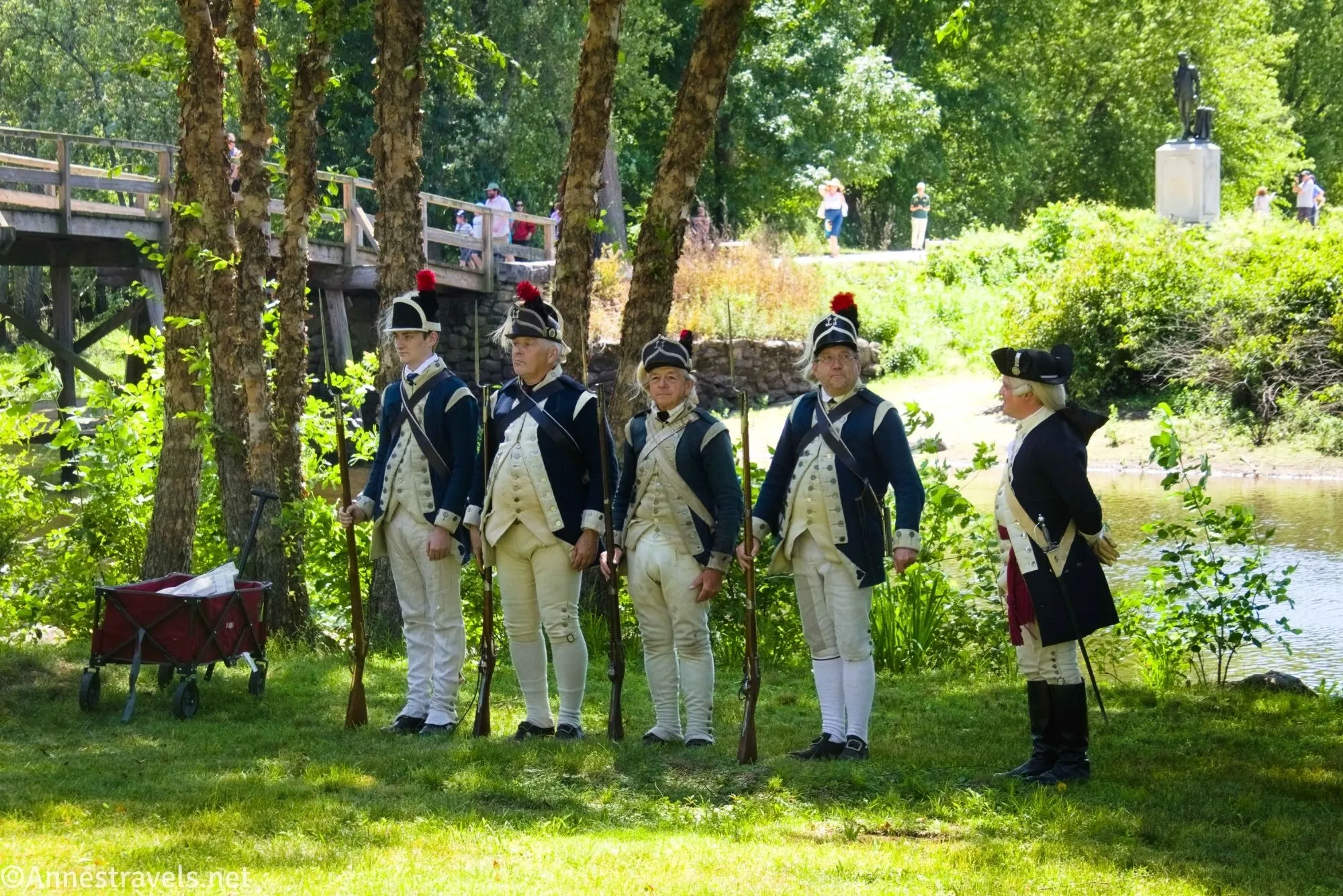 Five soldiers in costume of the Continental Army wait with their muskets beside a river with North Bridge in the background