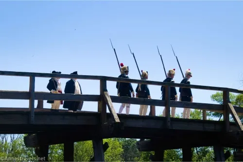 Four men with muskets march onto North Bridge while two other men look on