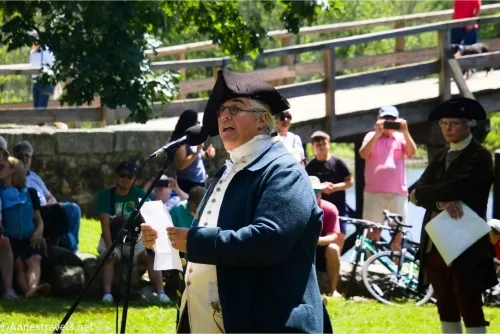 An older man in a costume from the Revolutionary War and a three-cornered hat speaks into a microphone while people watch