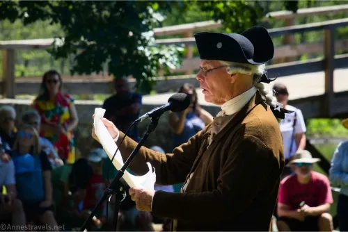 An old man in a brown jacket and black three-cornered hat reads from old-looking papers into a microphone with North Bridge in the background
