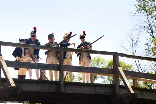 Four men in old soldier's uniforms from the Revolutionary war stand on a bridge loading their muskets
