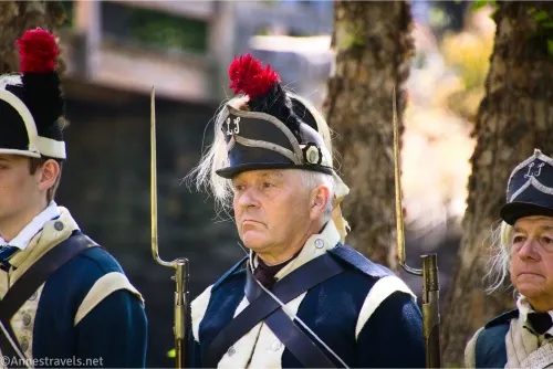 A solder with a dark blue and white uniform and fancy hat complete with a red feather holds a musket with a bayonet