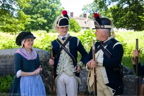 A woman in historic costume smiles at two men dressed as though they were in the Continental Army with The Manse in the background across a green field