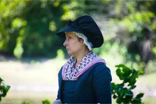 A woman wearing Revolutionary War-era clothing, including a black bonnet, pink shawl, and a dark blue dress