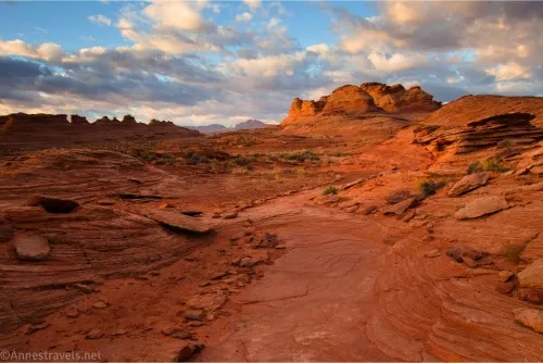 A slickrock path travels toward rock formations while the sun rises on clouds in the sky