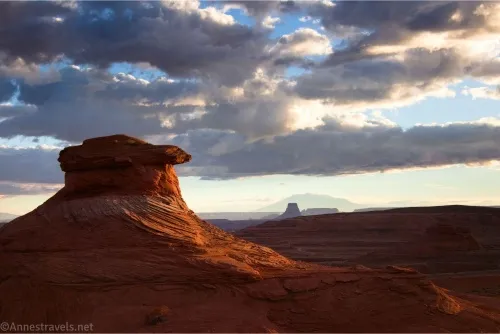 A single rock formation outlined on one side by the sun with clouds above and a silhouette of a mountain and a rock formation in the distance