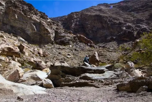 A hiker has successfully traversed several boulders in a desert canyon with towering cliffs above 