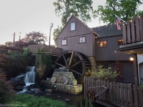 A brown building with a water wheel, several brown fences, an American flag, and a waterfall 