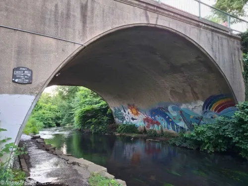 A concrete bridge over a stream and painted with trees, a compass, a sun, waves, a lobster, and a rainbow 