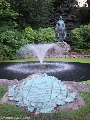 A corroded plaque is in front of a fountain ringed with duckweed and a statue of a woman and trees