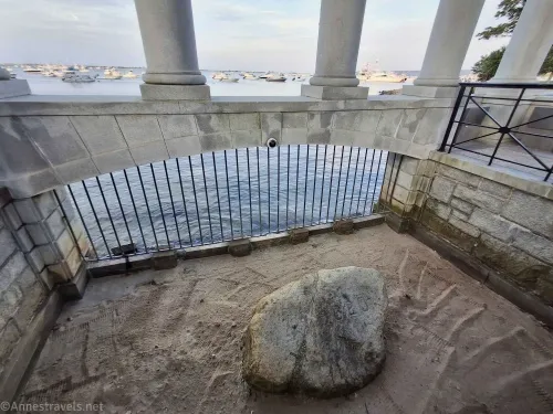 A rock sits on sand with a metal grate and pillars overlooking moored boats in the harbor 