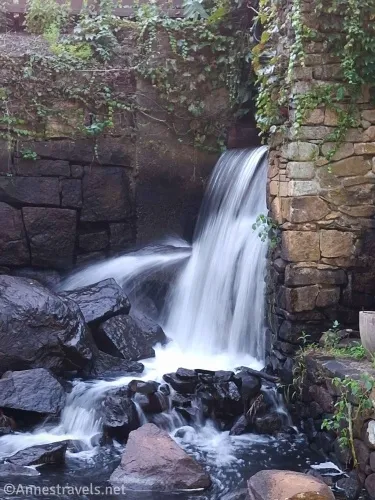 A waterfall tumbles from behind a stone wall with ivy onto black rocks 