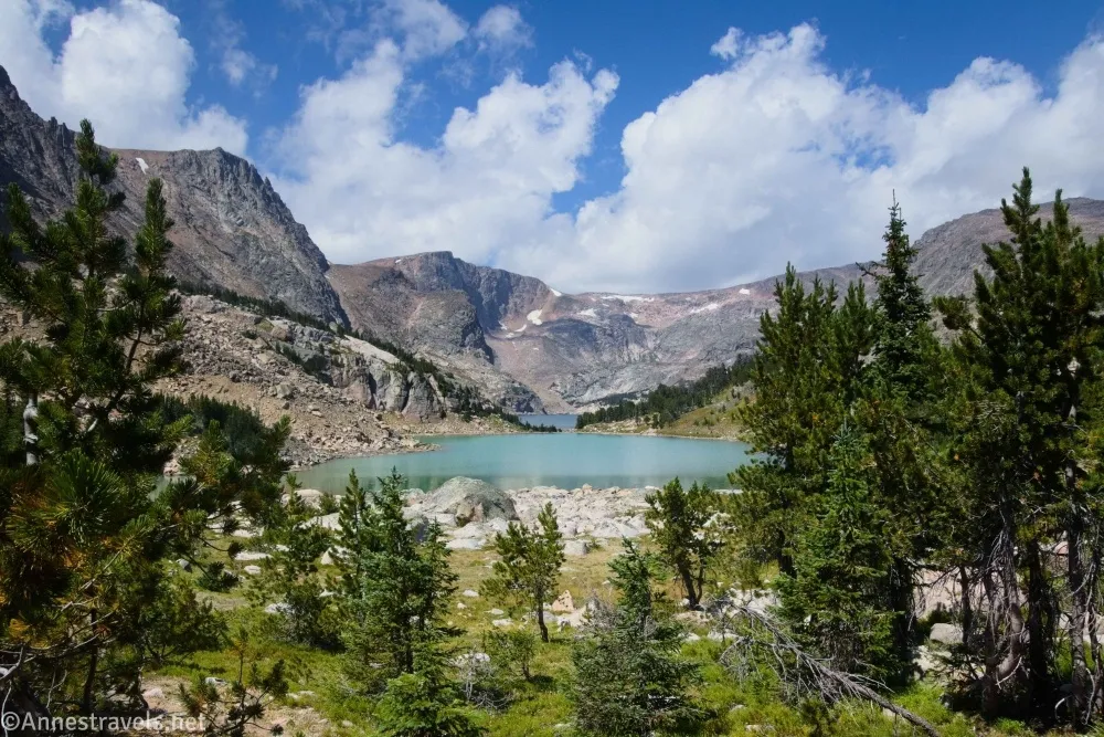 Green fir trees frame a green-blue lake and towering gray mountains below white puffy clouds and a blue sky