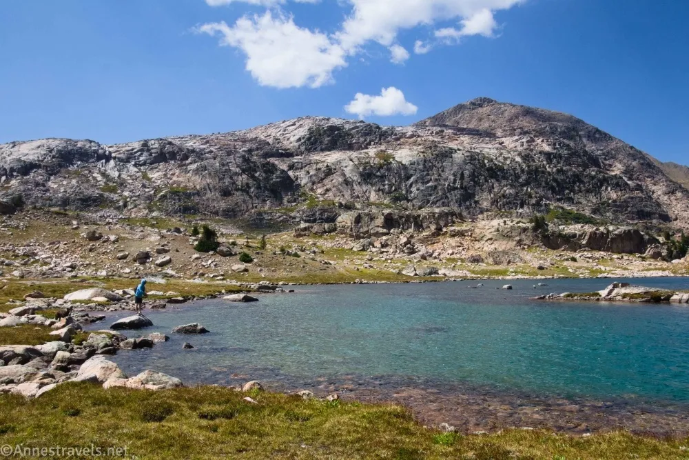 A man in a blue sun shirt fishes in a blue lake below the gray cliffs of mountains