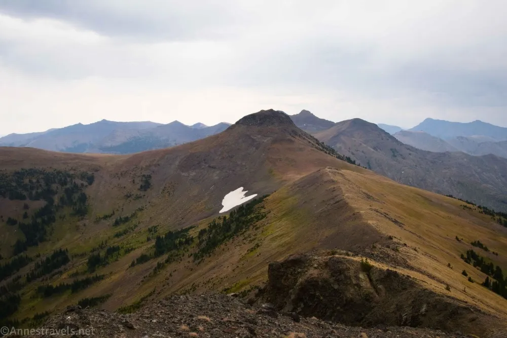 Meadow-covered mountains rise in front of gray cliffy mountains below a dark and threatening cloudy sky