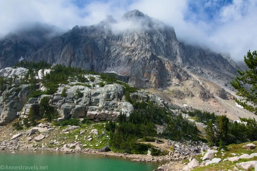 Clouds around the tops of gray cliffy mountains with the corner of a green lake below