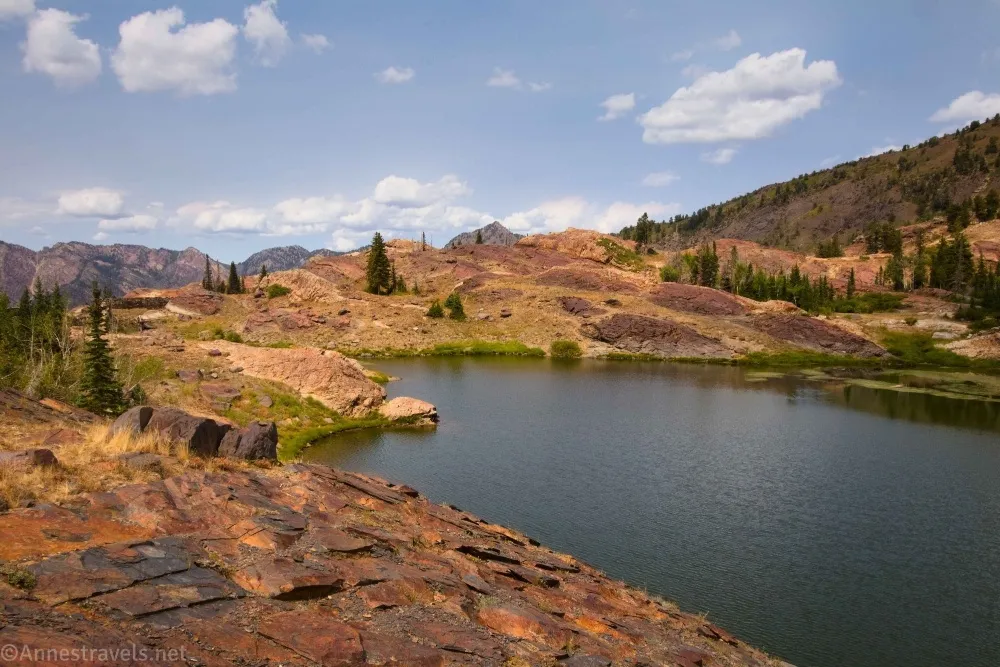 Puffy clouds sail above red rocks and mountains above a small lake