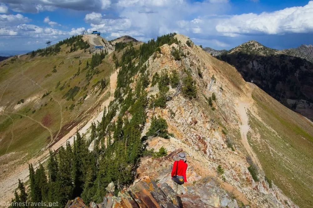 A hiker in a red shirt stands on a rocky ridgeline looking at other mountains and clouds in a blue sky