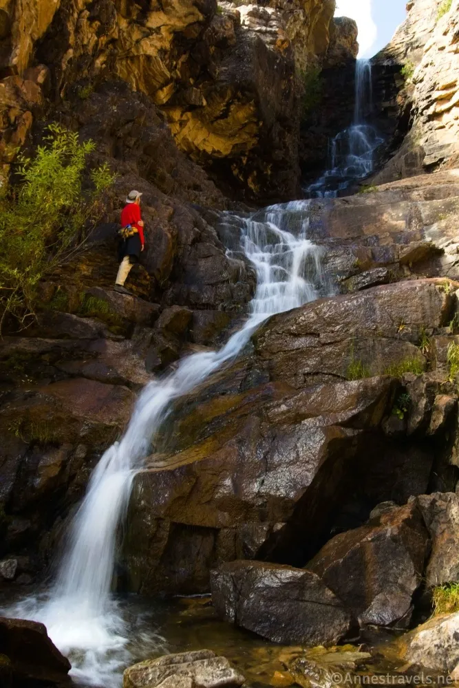 A hiker in a red shirt stands to the left of a very tall waterfall running down wet rocks