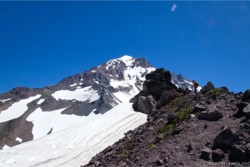 A hiker stands on a rocky ridge with snow and the peak of Mount Hood beyond