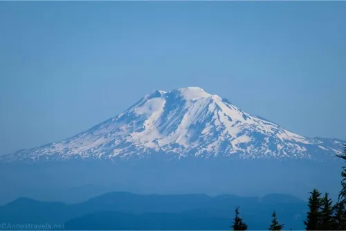 A snowy, hazy volcano rises out of blue hills into a blue sky