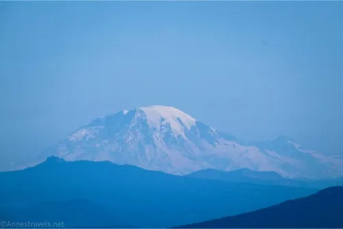 A hazy, snowy rounded volcano rises from blue mountains 