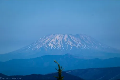 A hazy volcano with a smattering of snow and a flat or missing top rises above blue hills and sits below blue skies