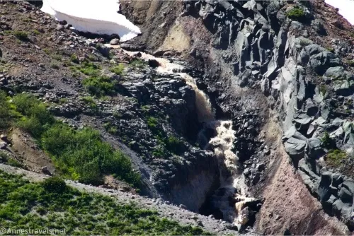 A muddy river flows from under a patch of snow between rocky banks